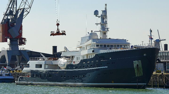 Dark-hulled explorer yacht with a white superstructure moored in a harbour beside a crane